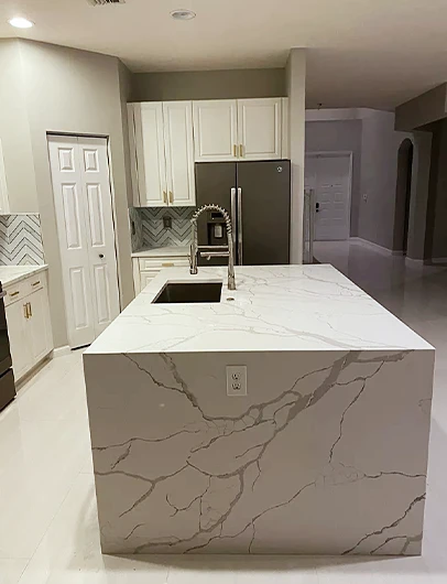 A modern kitchen with a large white marble-patterned island, white cabinets, and a dark refrigerator.