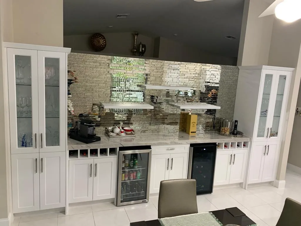 A modern home bar with white cabinets, two built-in beverage refrigerators, and a unique mirrored tile backsplash.