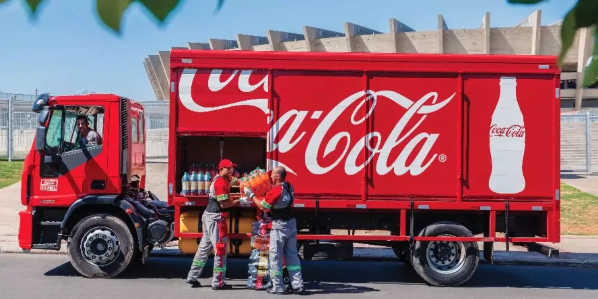 A foto mostra um caminhão da Coca-Cola e dois ajudantes